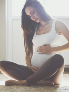 Pregnant woman sitting cross legged on the floor looking down and placing a hand on her baby bump. Reflecting and grouding with breath